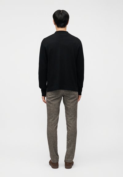 Άνδρας με την πλάτη στραμμένη, φορώντας μαύρο μακρυμάνικο πουκάμισο, γκρι υφασμένα παντελόνια και καφέ παπούτσια, στέκεται μπροστά σε απλό ανοιχτόχρωμο φόντο.