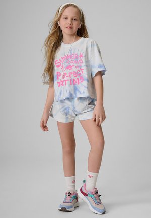 Młoda dziewczyna z długimi włosami, nosząca słuchawki, krótkie spodenki w stylu tie-dye i t-shirt z różowym napisem, białe skarpetki i kolorowe trampki, stojąca na szarym tle.