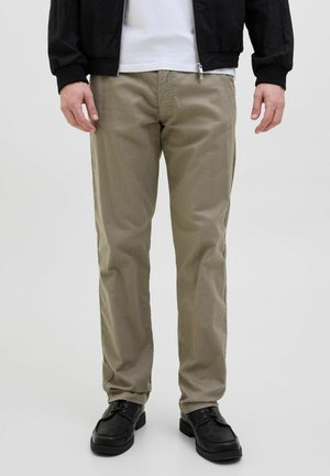 Beige Baumwollhosen mit geradem Schnitt und subtiler Textur. Getragen mit schwarzen Schuhen und einer schwarzen Jacke über einem weißen Hemd.