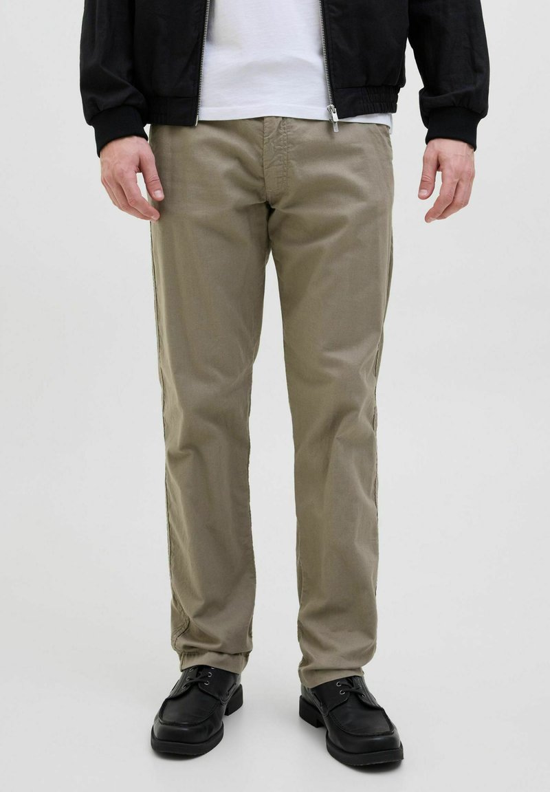 Beige cotton trousers with a straight fit and subtle texture. Worn with black shoes and a black jacket over a white shirt.