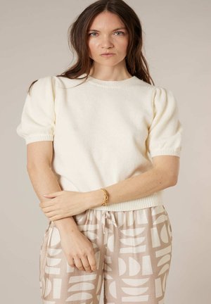 Femme aux longs cheveux bruns portant un pull blanc à manches bouffantes et un pantalon beige avec des motifs géométriques blancs abstraits, debout les bras croisés.