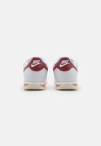 Hvite sneakers med røde detaljer, med en teksturert hæl, kontrastlogo og en tykk såle med et mønstrete dekk for grep og stabilitet.