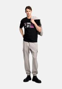 Camiseta negra con estampados gráficos en blanco y púrpura, combinada con pantalones cargo beige con bolsillos laterales. El modelo sostiene una sudadera gris.