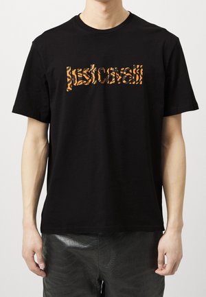 Person trägt ein schwarzes Kurzarm-T-Shirt mit orangefarbenem, gemustertem Text auf der Brust und schwarze, strukturierte Hose.