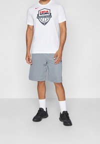 Bílý Nike tričko s grafikou "USA BASKETBALL", spárované se šedými sportovními kraťasy. Nosené s bílými ponožkami a černými teniskami.