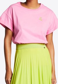 Kvinna i en lös rosa T-shirt med kort ärm som har en liten grön broderad design, tillsammans med en högmidjad limegrön plisserad kjol.