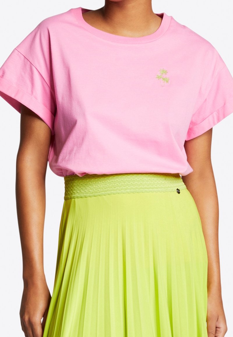 Kvinna i en lös rosa T-shirt med kort ärm som har en liten grön broderad design, tillsammans med en högmidjad limegrön plisserad kjol.