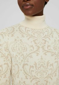 Närbild av en person som bär en krämfärgad turtleneck-tröja med ett beige, intrikat blommönster och paisleymönster.