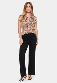 Blumenmusterte, gerüschte Bluse in Beige- und Orangetönen, kombiniert mit schwarzen, weit geschnittenen Hosen. Mit Riemchensandalen in Schwarz wird das Outfit abgerundet.