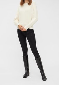Kremowy sweter z prążkowanym dołem, czarne rurki i czarne skórzane wysokie botki. Neutralna paleta kolorów, gładka faktura, elegancki design.
