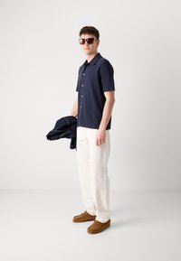 Camicia corta blu navy con bottoni, pantaloni crema, scarpe mocassino in suede marroni. Modello che tiene in mano un giubbotto in denim scuro, indossando occhiali da sole.
