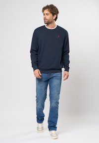 Hanorac bleumarin cu guler rotund și manșete din tricot, având un mic logo roșu pe piept. Asortat cu blugi bleu deschis și adidași de culori deschise.