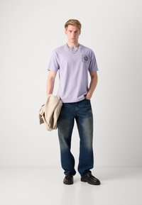 Lys lilla bomull T-skjorte med et sirkulært grafisk trykk, kombinert med blå denimjeans og svarte sko, holder en beige jakke.