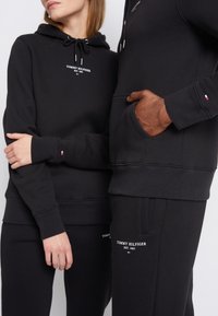 Czarne bluzy z kapturem z białym tekstem logo, pasujące czarne spodnie dresowe. Oba elementy odzieży mają minimalistyczny design, z kieszeniami i markowymi akcentami.