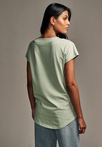 Hellgrünes, kurzärmliges T-Shirt mit Rundhalsausschnitt und lockerer Passform, mit abgerundetem Saum, aus weichem Baumwollstoff, getragen mit hellblauer Jeans.
