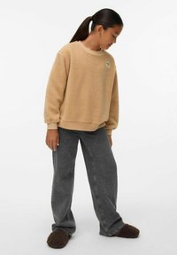 Béžová fleecová mikina s kulatým výstřihem, dlouhými rukávy a žebrovanými manžetami, spárovaná s tmavě šedými širokými džíny a hnědými chlupatými pantoflemi.