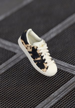 Schwarzer und beiger Wildledersneaker mit drei schwarzen Streifen, weißen Schnürsenkeln und einer Schalenspitze. Verfügt über eine strukturiere Sohle und ein goldenes Logo an der Ferse.