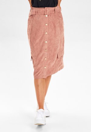 Vrouw draagt een roze corduroy midi rok met knopen aan de voorkant en een riem, gecombineerd met witte platformsneakers.
