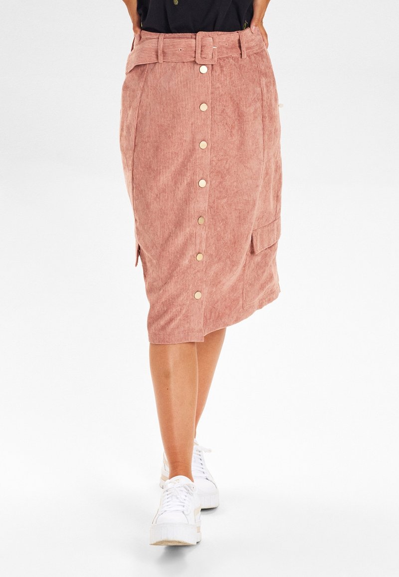 Femme portant une jupe midi en velours côtelé rose avec des boutons sur le devant et une ceinture, assortie à des baskets blanches à plateforme.