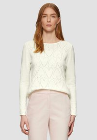 Pull en maille blanc avec motif en cœur, col rond, manches longues et texture douce, associé à un pantalon tailleur rose clair.