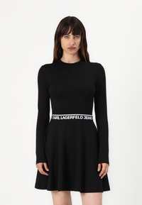 Black long-sleeve dress with a flared skirt, featuring a white branded waistband. Smooth fabric texture, fitted bodice design.