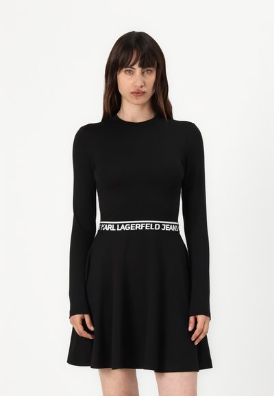 Robe noire à manches longues avec une jupe évasée, dotée d'une ceinture de marque blanche. Texture de tissu lisse, design de corps ajusté.