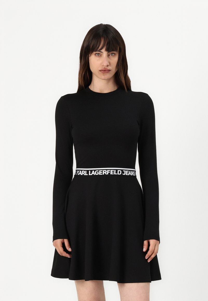 Black long-sleeve dress with a flared skirt, featuring a white branded waistband. Smooth fabric texture, fitted bodice design.