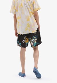 Camicetta a maniche corte tie-dye bianca e gialla abbinata a pantaloni corti neri con fiori. Calzature blu forate completano l'outfit.