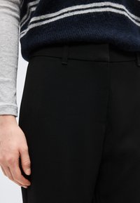 Άτομο που φοράει μαύρο παντελόνι ραμμένο στα μέτρα και μπλε σκούρο πουλόβερ με λευκές ρίγες, μερικώς εμφανές τον κορμό και το αριστερό χέρι χαλαρό στο πλάι.