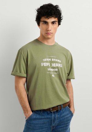 Giovane uomo con capelli scuri e ricci, che indossa una t-shirt verde Pepi Jeans London e jeans blu con le mani nelle tasche, su uno sfondo semplice.