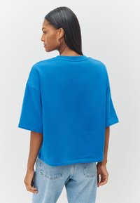 Zila oversized kokvilnas T-krekls ar apaļu kakla izgriezumu un īsām piedurknēm, kopā ar gaiši izbalējušām džinsu biksēm. Gluda tekstūra. Nav redzamu rakstu.