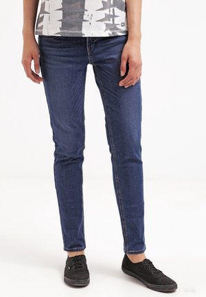 Personne portant un jean slim bleu foncé, des baskets noires et une chemise à manches courtes à motifs, debout sur un fond blanc.