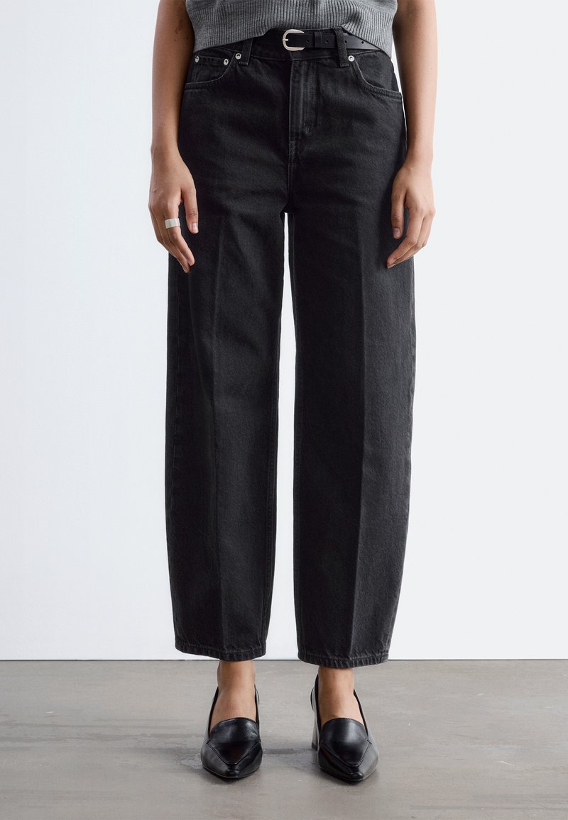 Person som bär svarta raka jeans med hög midja och svart bälte, grå topp, svarta spetsiga skor, står på ett grått golv.