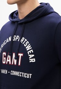 Granatowy hoodie wykonany z mieszanki bawełny, z kapturem na sznurki oraz wyraźnym białym/czerwonym tekstem z logo "American Sportswear" i "GANT".