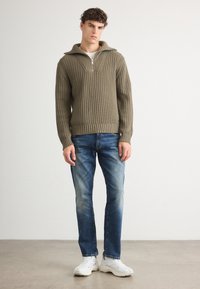 Maglione a coste verde oliva con collo a mezza zip, abbinato a jeans blu strappati e sneaker bianche, in piedi su una superficie liscia.