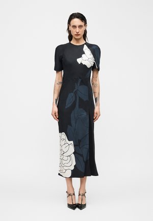 Frau mit dunklen Haaren, die ein schwarzes Midikleid mit großen weißen und marineblauen Blumenmustern, kurzen Ärmeln und schwarzen spitzen Absätzen trägt.