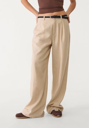 Pantalons larges beiges avec des plis à l'avant, confectionnés dans un tissu léger. Associés à une ceinture tressée noire et des baskets marron.