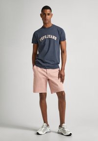 Jūrinės medvilninės marškinėliai su "Pepe Jeans" logo kremine spalva, derinami su šviesiai rožinėmis šortais. Modelis dėvi pilkus ir baltus sportbačius.