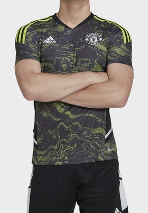 Mężczyzna ze skrzyżowanymi ramionami, ubrany w czarną i neonowo zieloną sportową koszulkę Adidas Manchester United oraz czarne spodenki.
