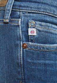 Blå denimjeans med en närbild på bakfickan. Har en vit etikett med röd "AG"-logotyp och en kopparknapp som accent. Lätt slitage synligt.