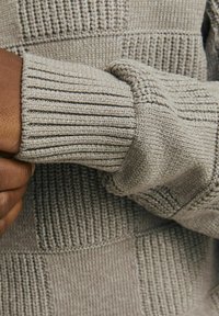 Sivi pleteni pulover s teksturiranim uzorcima, sa rebrastim rukavima i glatkim tijelom, naglašavajući varijacije u vrstama šavova.