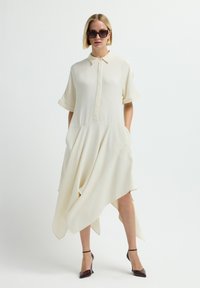 Vestido blanco asimétrico con cuello, mangas cortas y bolsillos laterales. Confeccionado en una tela suave y combinado con tacones altos oscuros.
