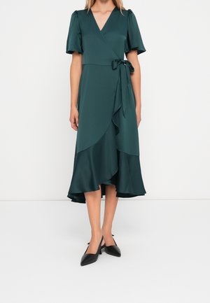 Kobieta w ciemnozielonej sukience kopertowej z falbankowymi rękawami i wiązaniem w talii, w połączeniu z czarnymi czółenkami slingback z ostrym noskiem.