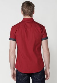Camisa roja de manga corta con cuello recto, puños oscuros de contraste azul marino y un pequeño logo en la parte posterior, combinada con jeans de mezclilla oscuros.