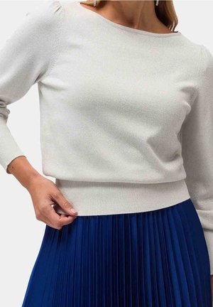 Kvinde iført en hvid langærmet top og et blåt plisseret skørt, der holder i kanten af toppen med den ene hånd.