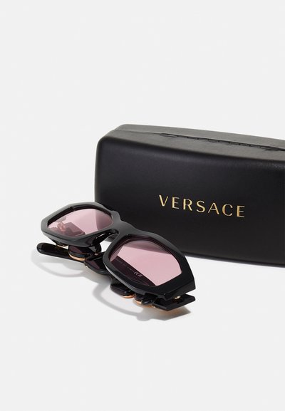 Sorte solbriller med pink linser, unik vinklet facon, guldfarvede detaljer og et Versace-logo på et sortetui.