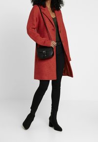 Cappotto rosso in misto lana con design sartoriale, maniche lunghe e bottoni frontali, abbinato a jeans skinny neri e stivaletti alla caviglia.