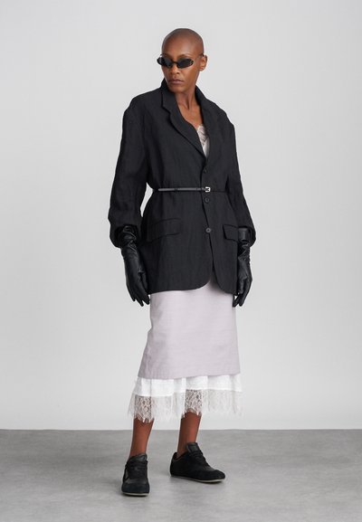 Crni laneni sako s skupljenim strukom, uparen sa svijetlo ljubičastom suknjom s čipkastim rubom i crnim cipelama. Model nosi crne rukavice i sunčane naočale.