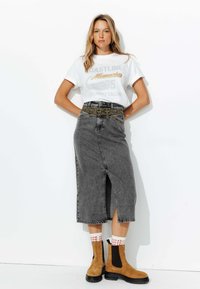 Jupe midi en denim gris avec une fente centrale, ceinture à motifs, t-shirt graphique blanc et bottines en daim marron à accents noirs.