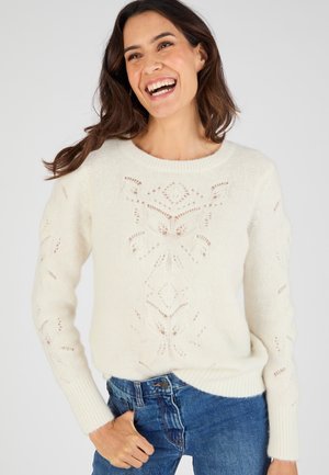 Pull crème en tricot présentant un motif texturé avec des détails ajourés sur les manches. Encolure ronde et poignets côtelés, associé à un jeans bleu.